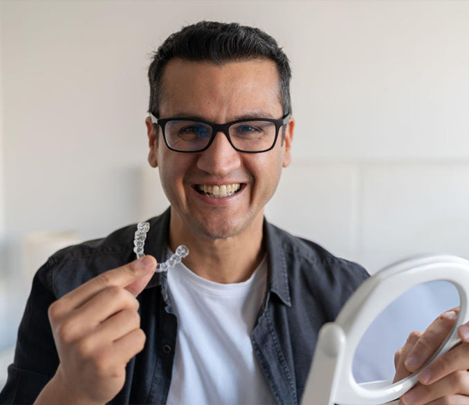 Man smiling at camera holding clear dental aligner and mirror in clinical setting.