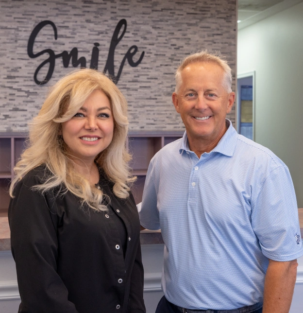 Two professionals posing together in orthodontic office with "Smile" sign visible on brick wall background.