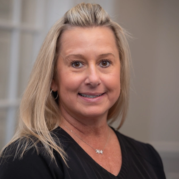 Professional woman with blonde hair smiling at camera in clinical office setting.