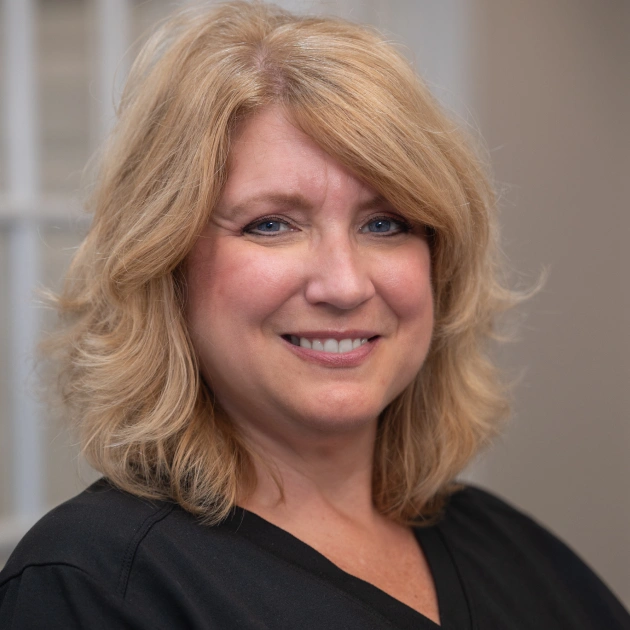 Professional woman smiling at camera in clinical office setting, wearing black top.
