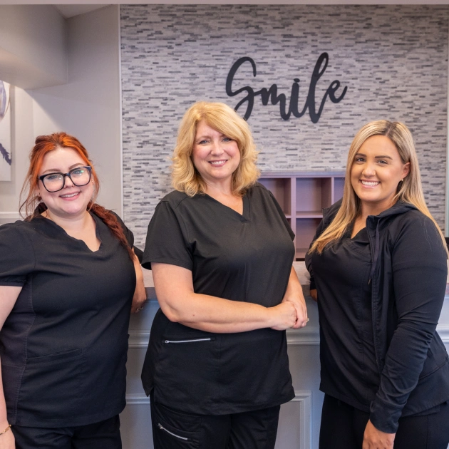 Three female dental staff members smiling in orthodontics office with "smile" wall sign.