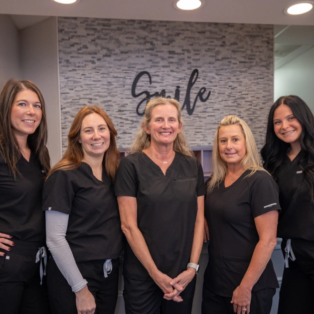 Five female dental professionals in black scrubs posing together in modern orthodontic office with "Smile" sign.