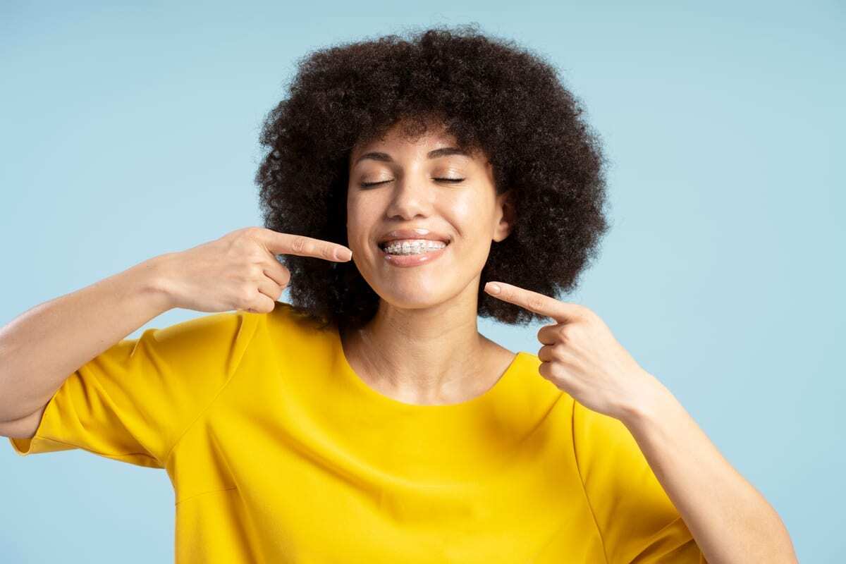 Young woman with braces smiling and pointing to her teeth against blue background.